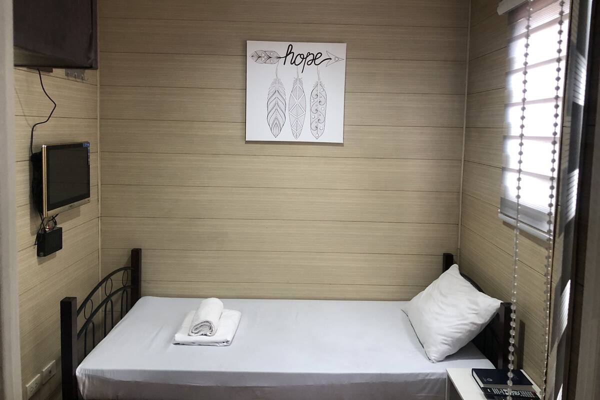 Een klein, netjes opgemaakt bed met een wit kussen en een opgerolde handdoek, een tv aan de muur en een kunstwerk met het woord 'hope' en veerdessins.