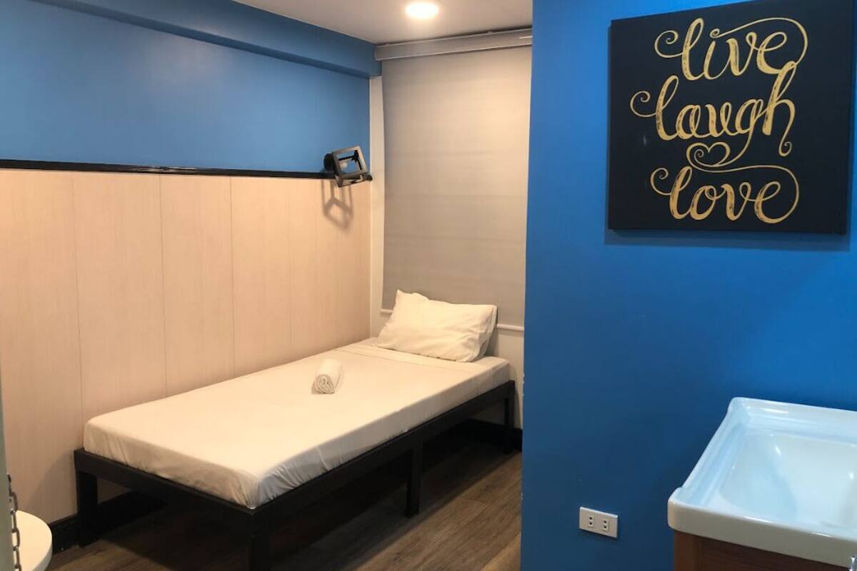 Een kleine eenpersoonskamer met een houten hoofdbord, een witte matras en een wandlamp. Op de muur staat een motiverende quote.