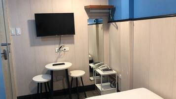 A hotel room with a bed, a TV mounted on the wall, a small table with two stools, and a mirror cabinet.