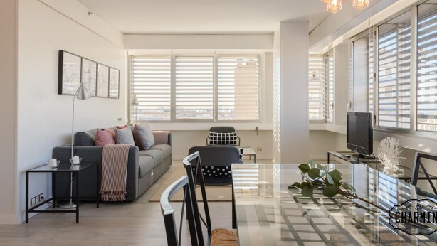 Una sala moderna con un sofá, una mesa de centro y un televisor. Cuenta con ventanas amplias con persianas, una araña y una planta sobre la mesa de centro.
