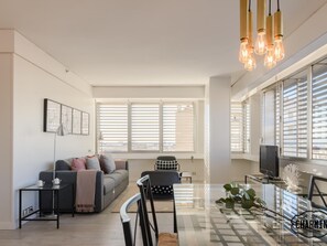 A modern living room with a sofa, a coffee table, and a TV. There are large windows with blinds, a chandelier, and a plant on the coffee table.