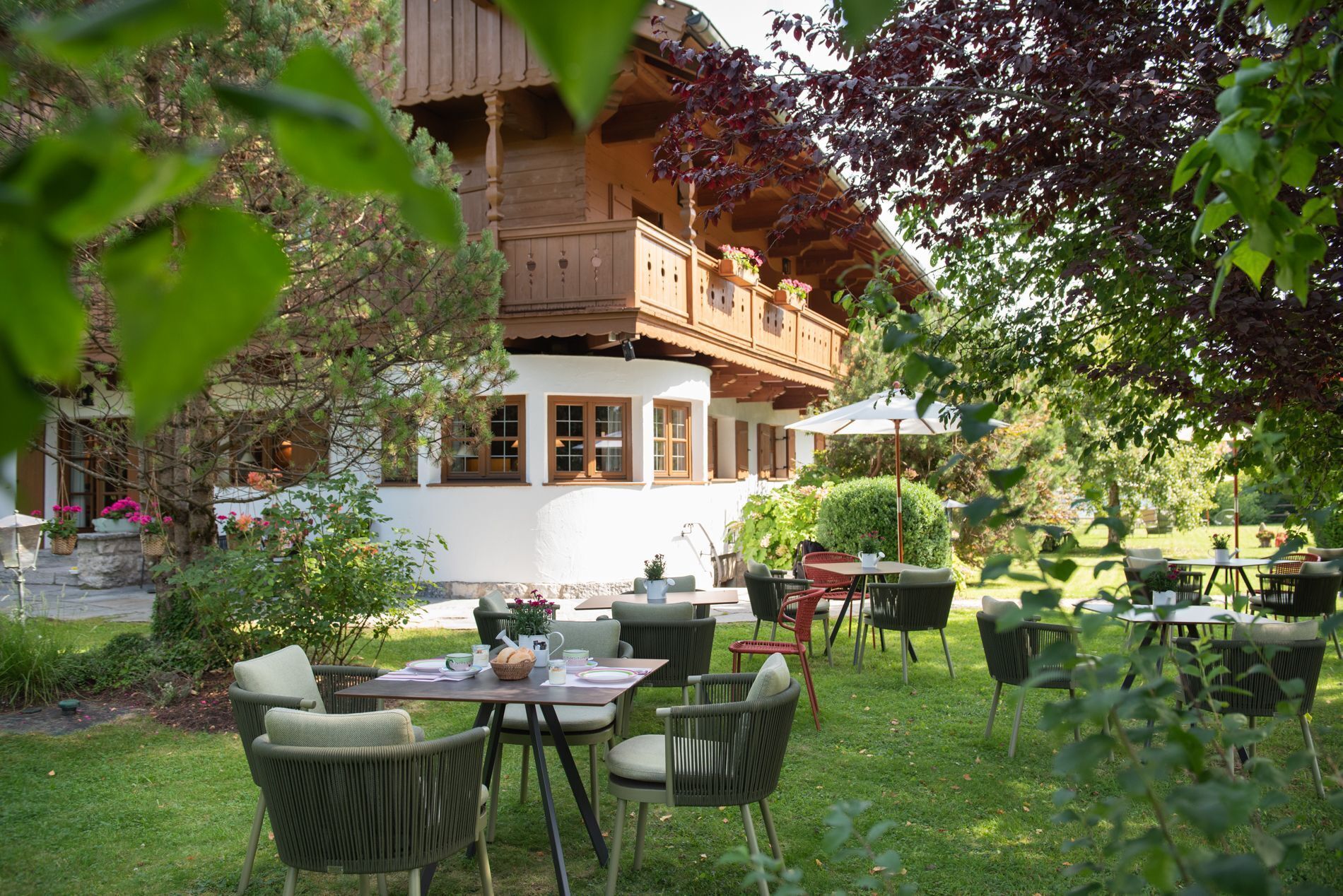 An outdoor seating area with wicker chairs and tables, surrounded by greenery and a wooden building with a balcony.