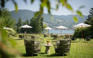 An outdoor seating area with wicker chairs and umbrellas, overlooking a lake and forested hills. - Single Standard - Landhaus Christl am See (Bad Wiessee)