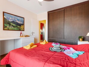 A bedroom with a red bedspread, a painting of a mountain landscape, a wardrobe, and a desk with a bottle of wine. - C103 big terrace, pool! (San Eugenio Alto)