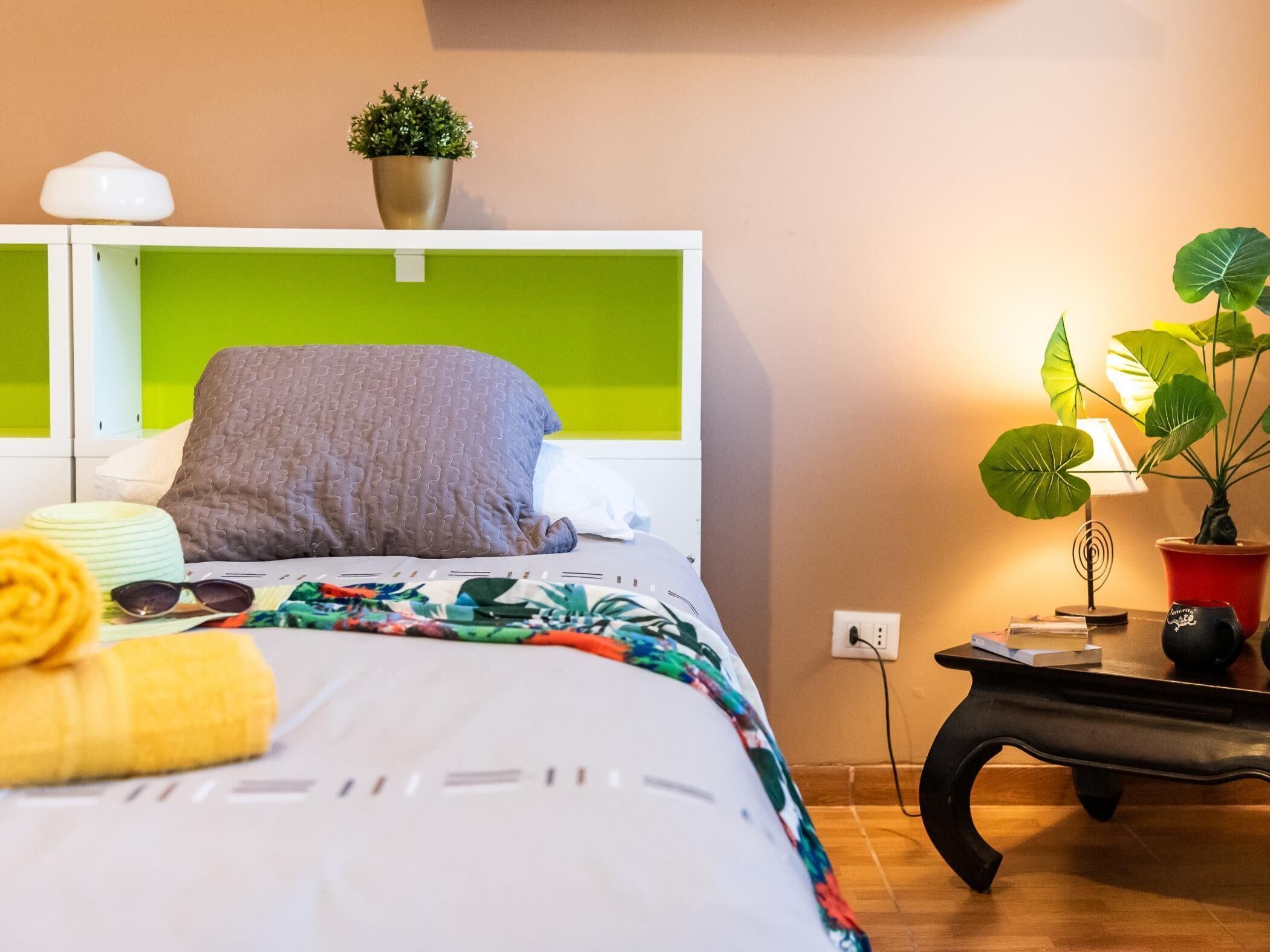 Un letto rifatto con una coperta a fiori, un cuscino grigio e un asciugamano giallo. C'è una piccola pianta sulla testiera, una lampada su un comodino e un vaso con una pianta al pavimento.