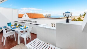 Un balcon avec une table dressée pour deux personnes, des chaises blanches, une plante en pot et une antenne parabolique.