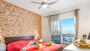 Une chambre d’hôtel moderne avec un mur décoré d’un motif en briques, un grand lit recouvert d’une couverture rouge, un ventilateur de plafond, un balcon avec vue et un plateau de petit-déjeuner sur la table.