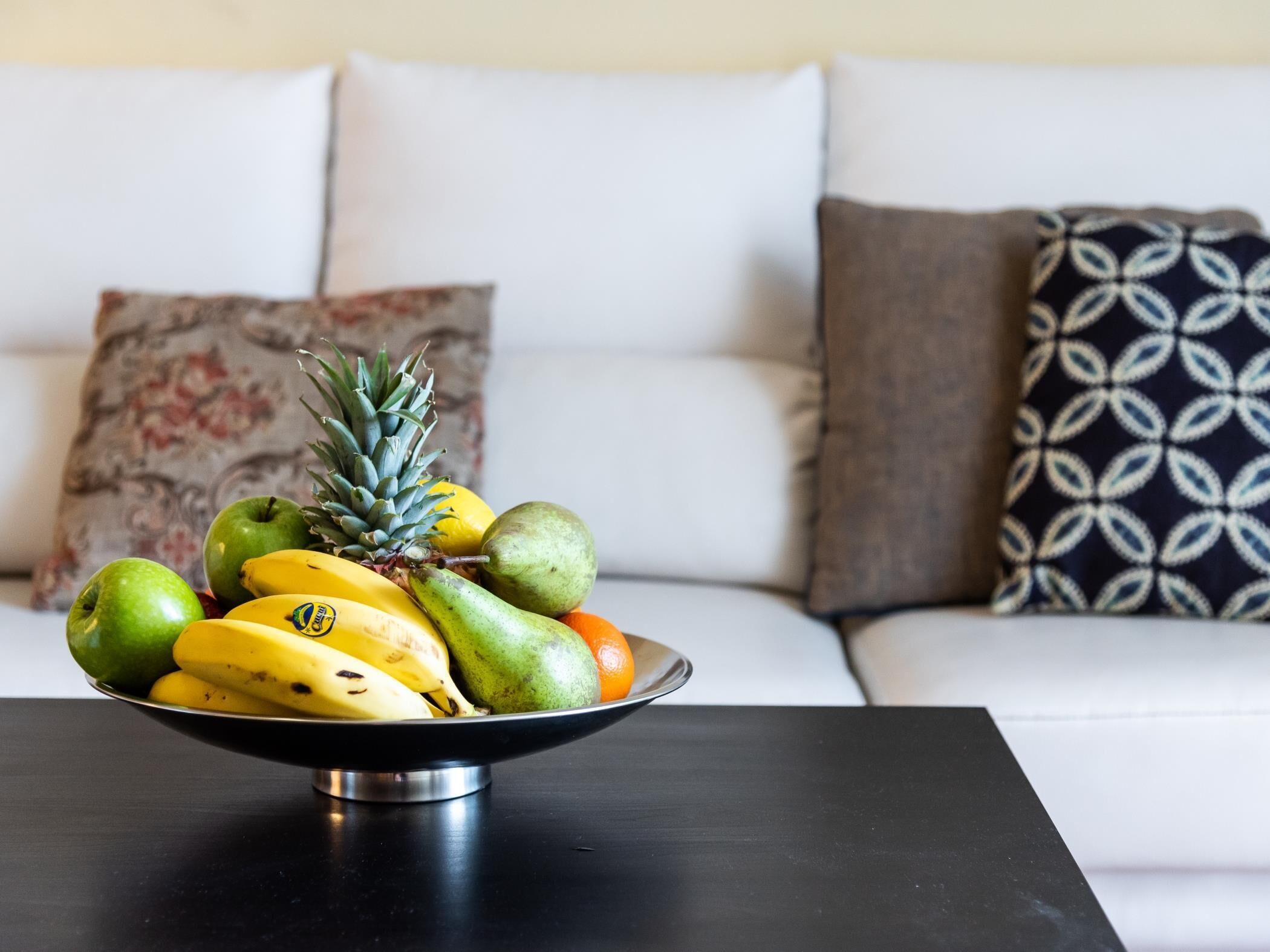 A fruit bowl on a table in front of a white sofa with patterned cushions.