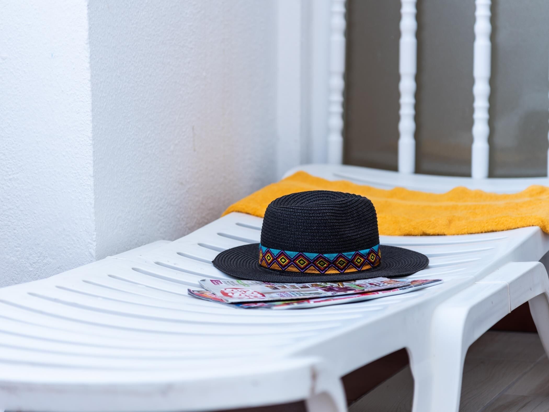 A white plastic chair with a black hat and a magazine on it, placed on a balcony with a yellow towel and a white wall in the background.