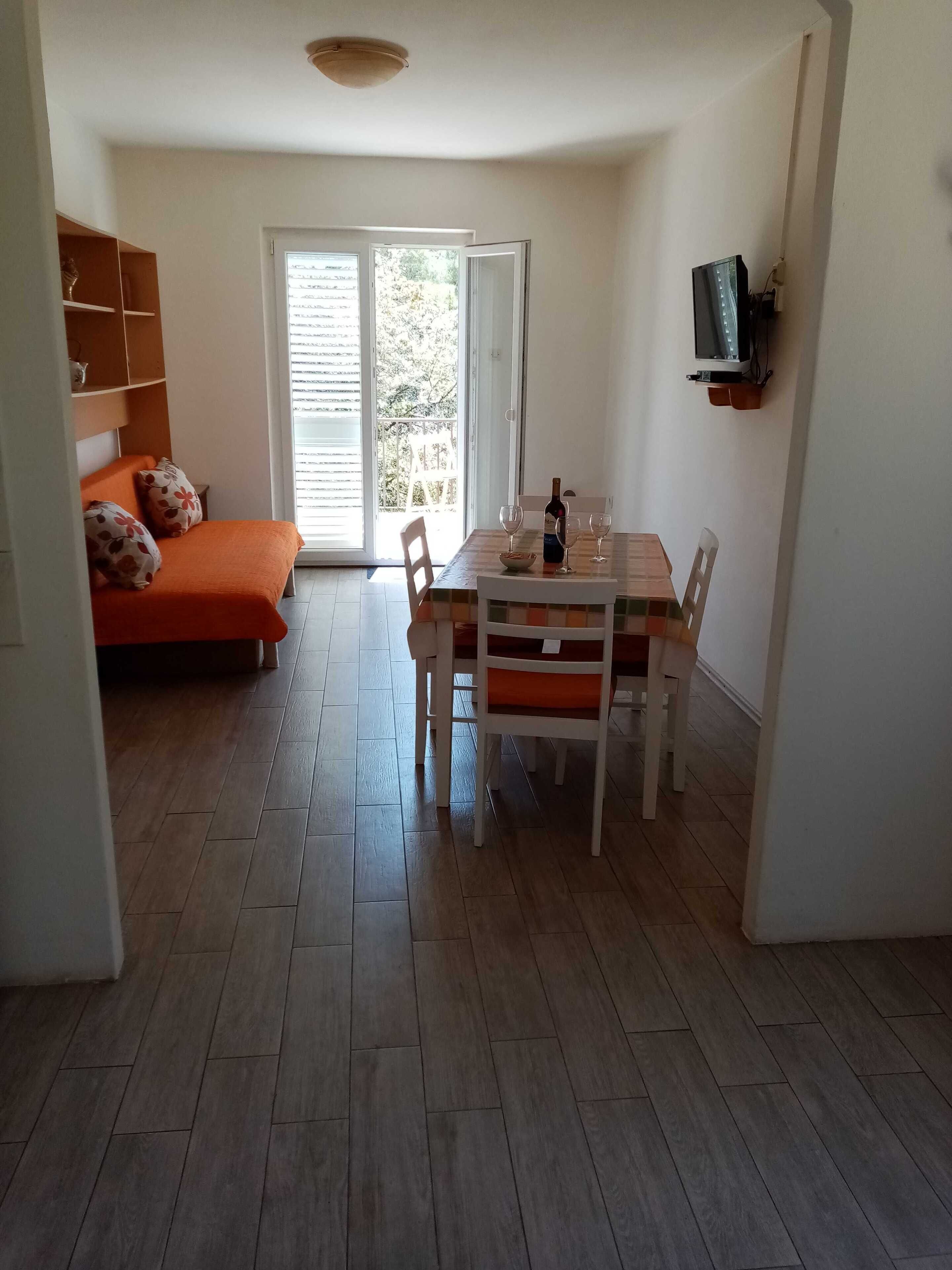 Una habitación con una mesa de comedor, sillas, un sofá con cojines florales, un televisor montado en la pared y un ventanal corredero que da a un espacio exterior.
