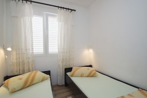 Quarto pequeno e limpo, com duas camas de solteiro, janela com cortinas brancas e chão de madeira.