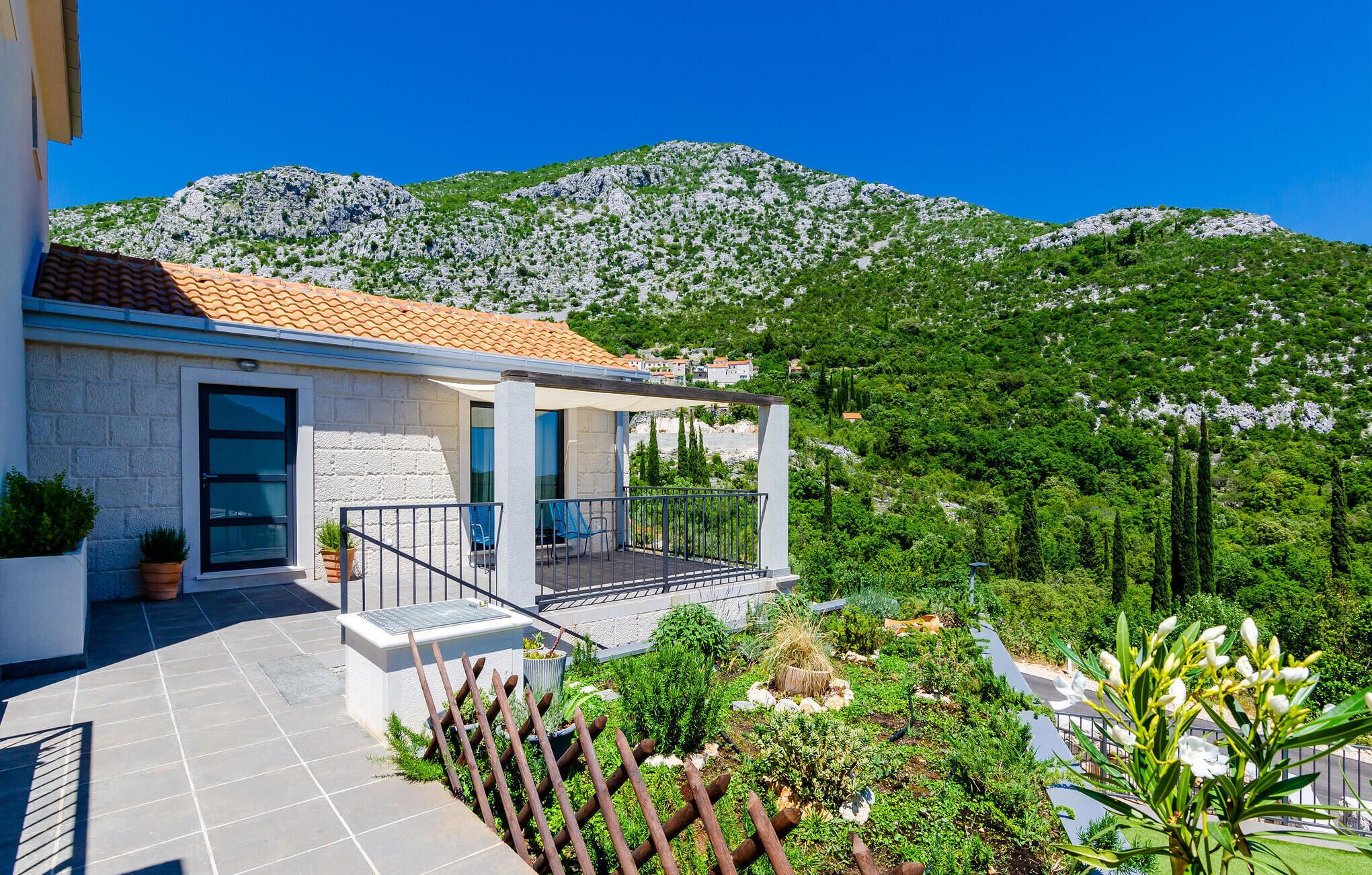Ein modernes Haus mit Ziegeldach, einem Balkon mit Metallgeländer und Blick auf grüne Hügel und Berge im Hintergrund.