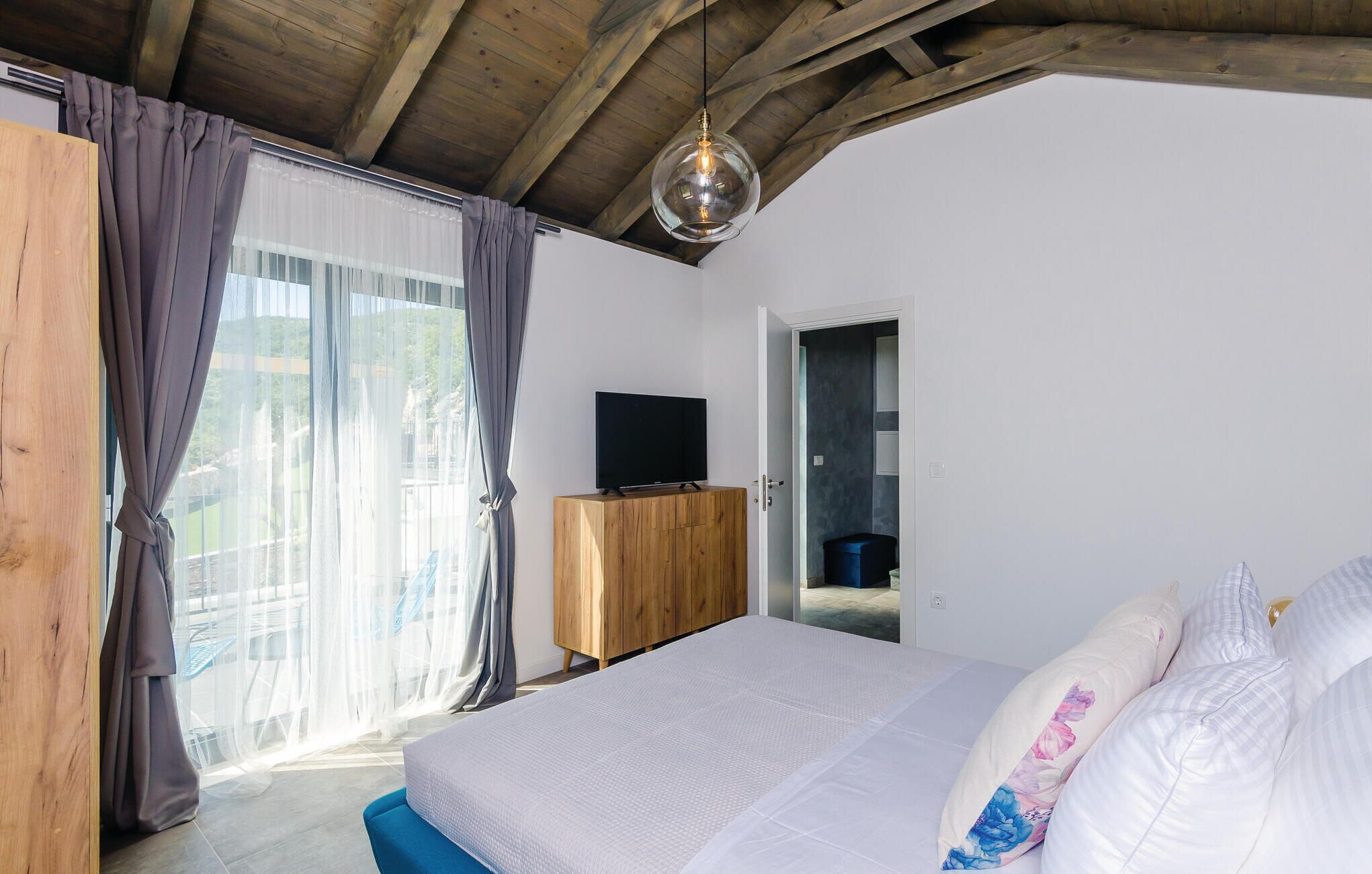 A bedroom with a bed, a wooden wardrobe, a television, and a view of the outdoors through a window with curtains.