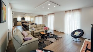 Ein modernes Wohnzimmer mit einer Sofagarnitur, einem Couchtisch und einem schwarzen Sessel. Im Hintergrund ist eine Küche mit Esstisch und Stühlen zu sehen.