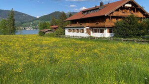 A traditional wooden chalet with a red-tiled roof, white walls, and wooden shutters, situated in a lush meadow with yellow flowers, overlooking a lake and mountains. - Double Deluxe - Landhaus Christl am See (Bad Wiessee)