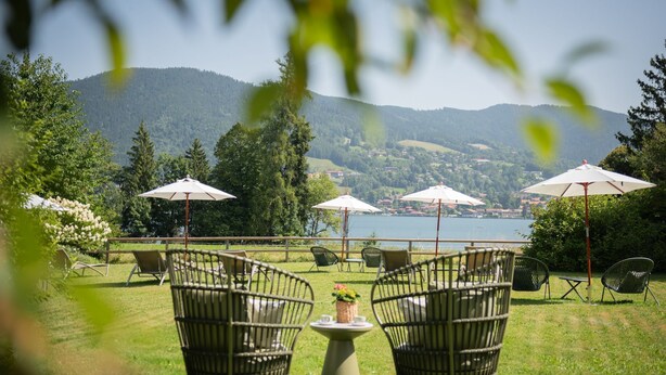 An outdoor seating area with wicker chairs and umbrellas, overlooking a lake and forested hills. - Double Deluxe - Landhaus Christl am See (Bad Wiessee)