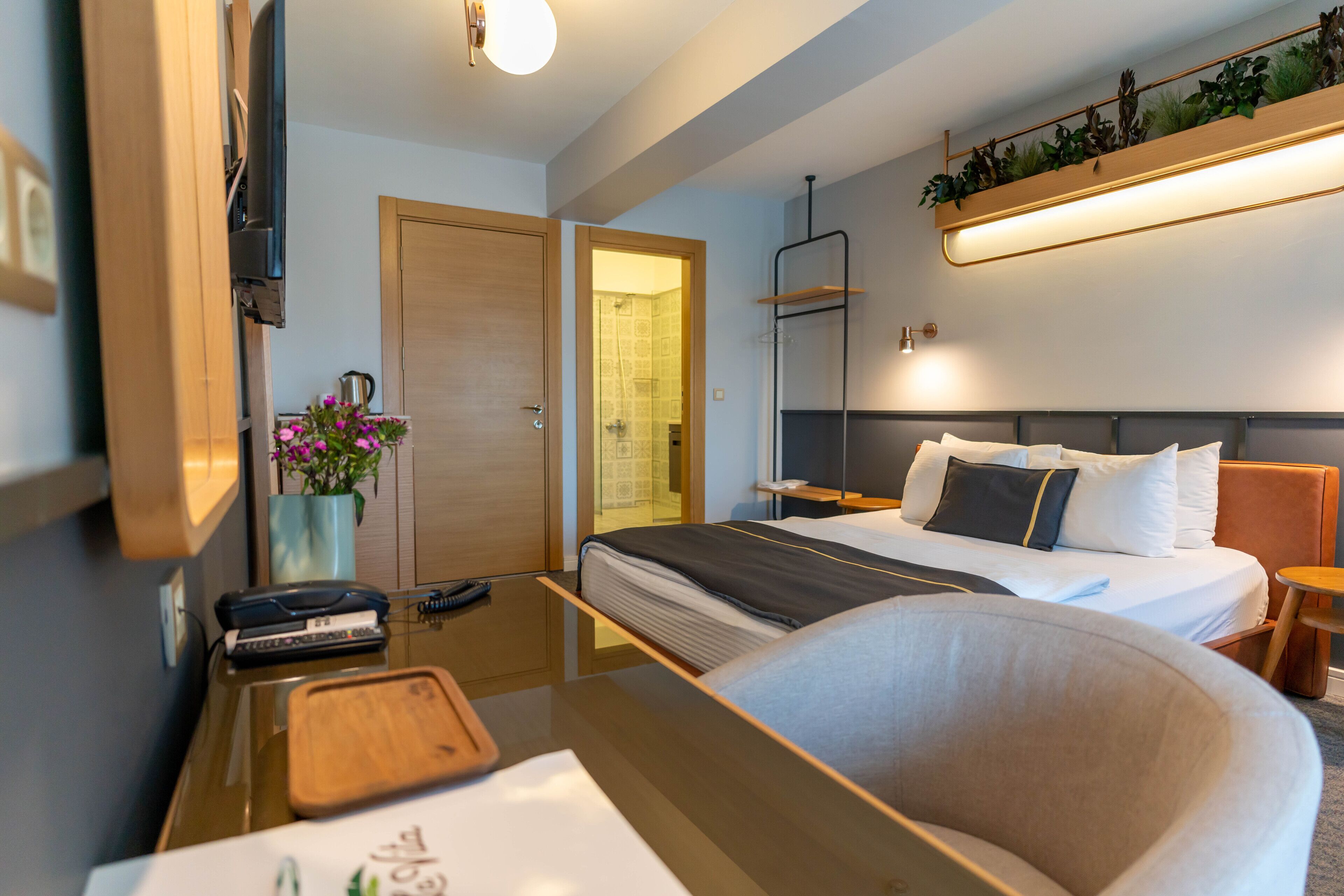 Una habitación de hotel moderna con cama, mesita de noche, silla y un pequeño escritorio con teclado y planta.