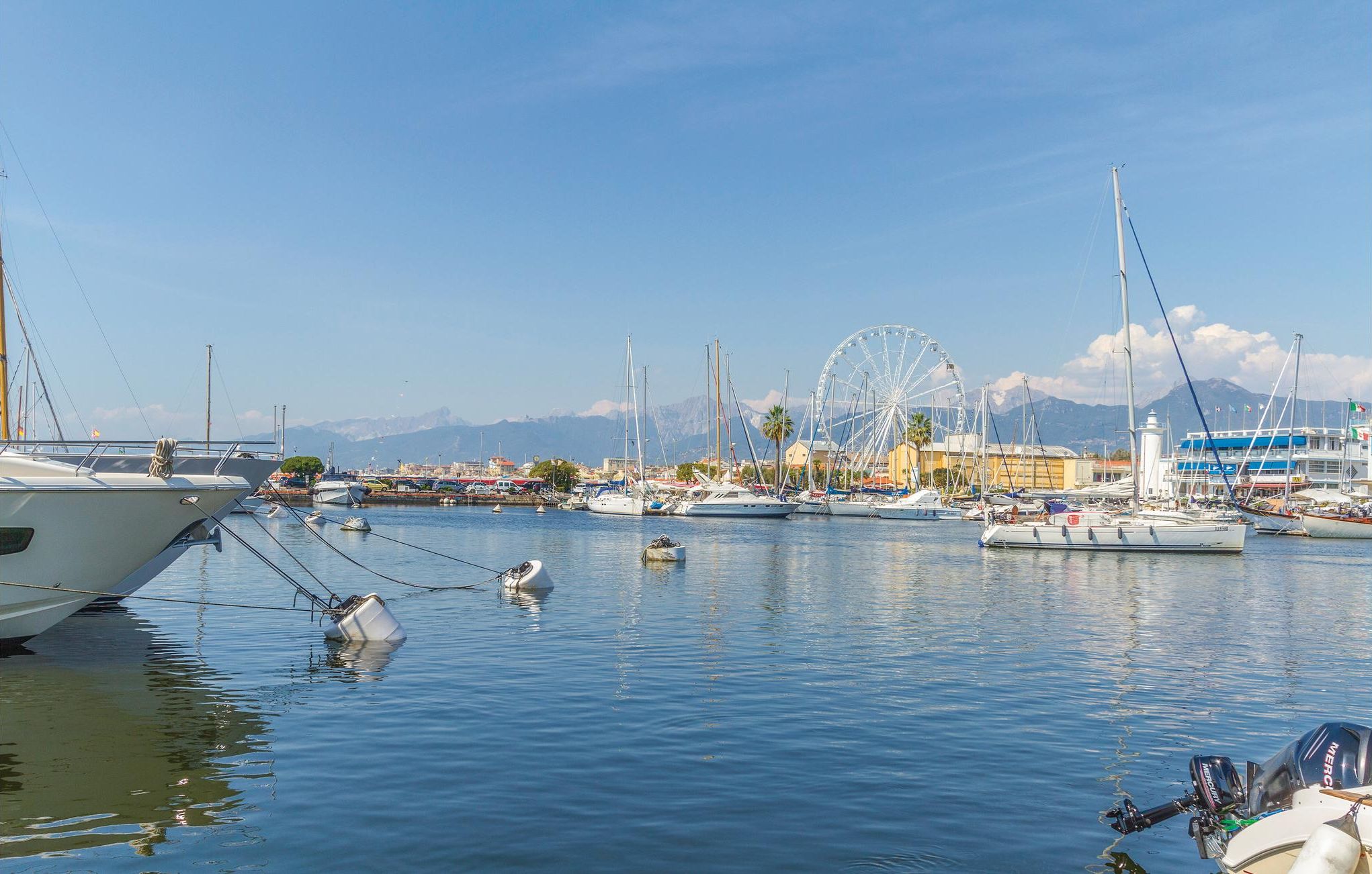 Un port de plaisance avec des bateaux amarrés, une grande roue et des montagnes en arrière-plan.