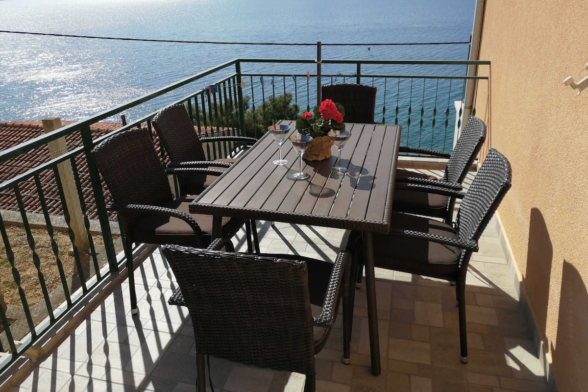A balcony with a table and chairs, overlooking a body of water and distant mountains.