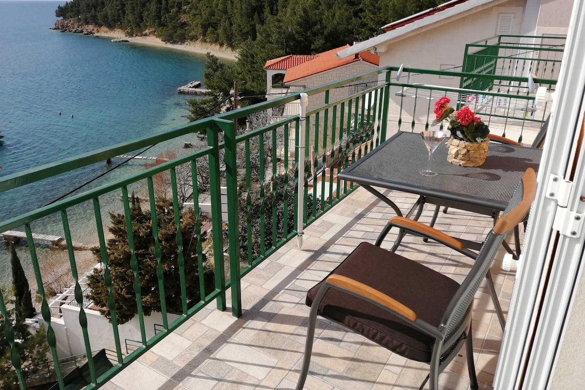 A balcony with a table and chairs, overlooking a beach and the sea.