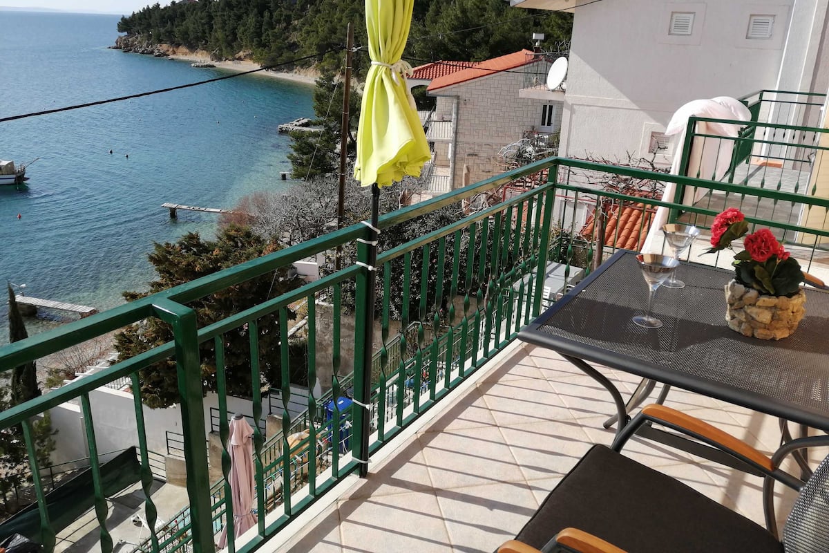 A balcony with a view of the sea, a table with chairs, and a potted plant.