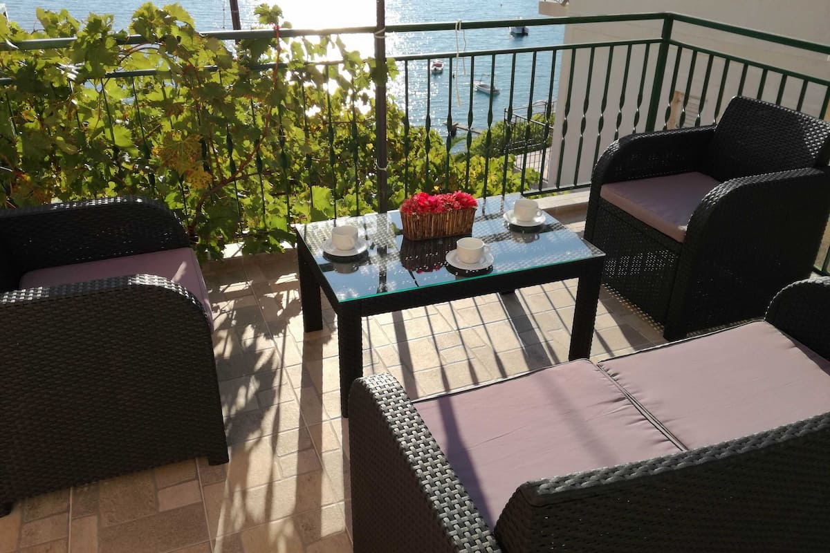 A balcony with wicker furniture, a glass table with cups and a basket of flowers, and a view of the sea.