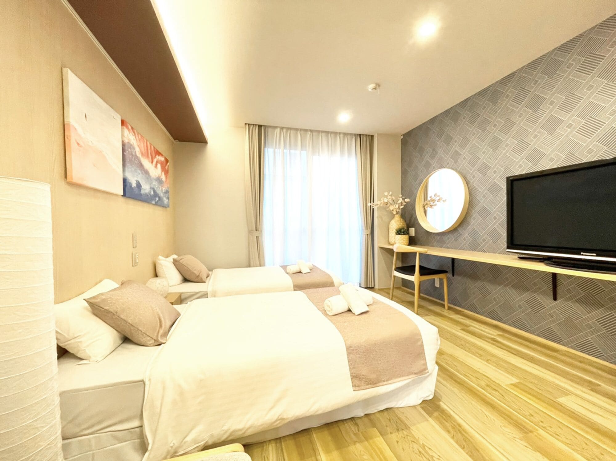 A hotel room with two beds, a TV, a desk, and a large window with curtains.