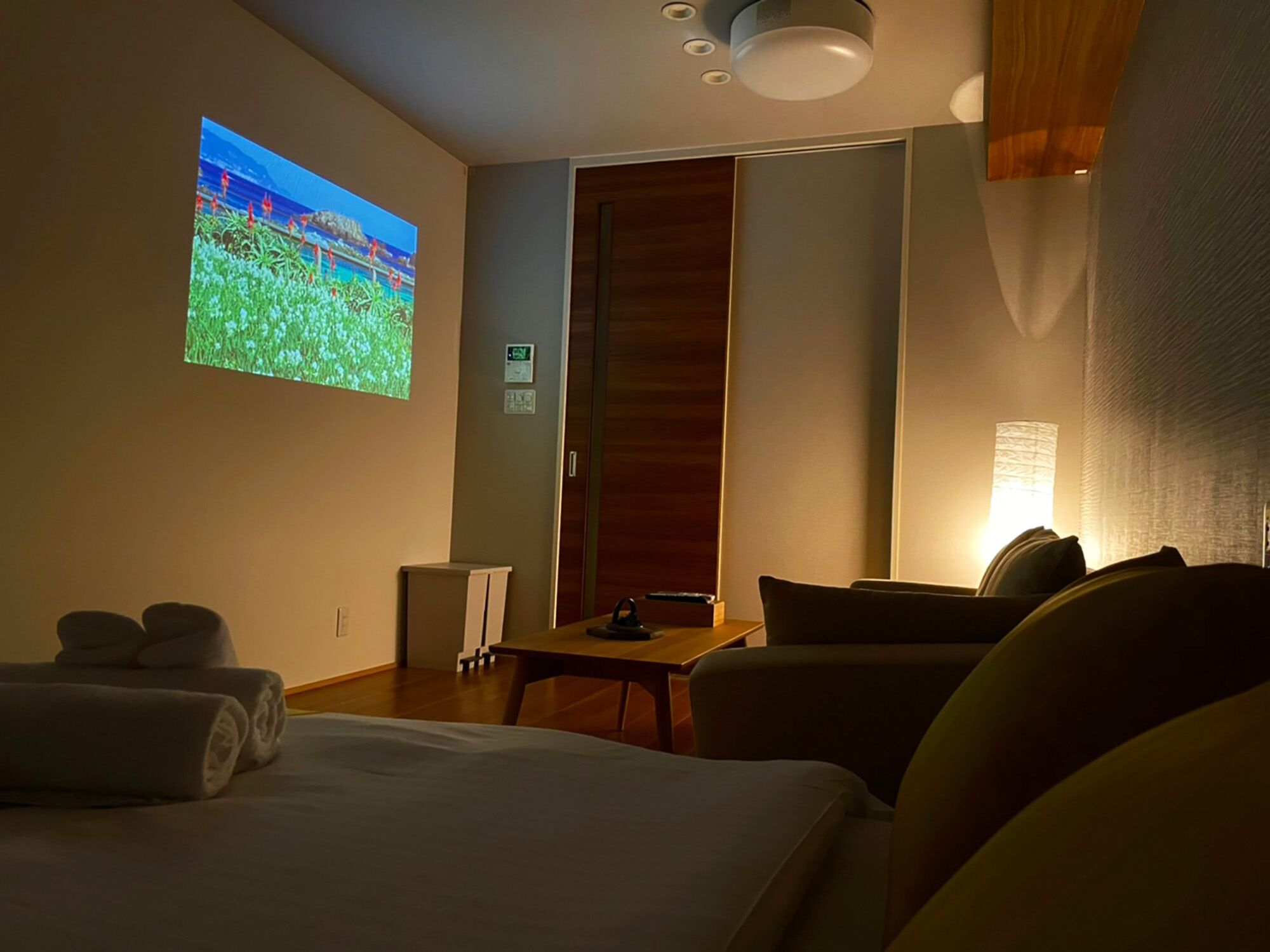 A hotel room with a bed, a desk, a chair, a TV screen, and a lamp.