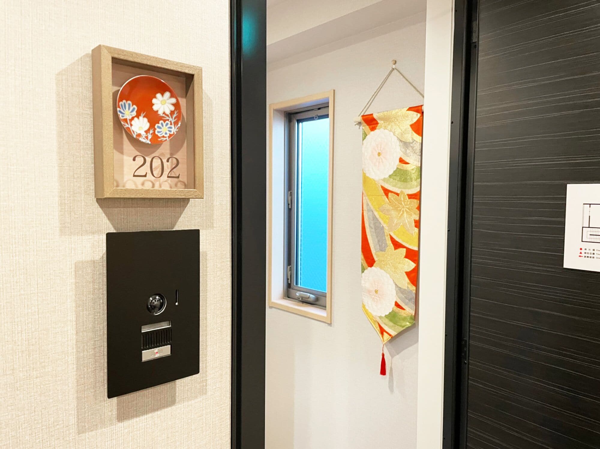 A wall with a framed number '202', a black intercom, and a decorative hanging with floral patterns.