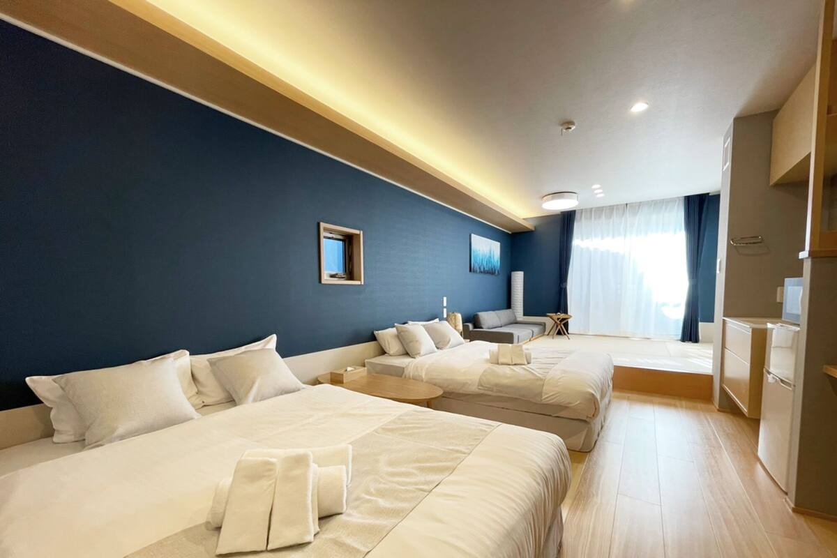 Habitación de hotel con tres camas, un pequeño rincón de descanso y vistas al exterior.