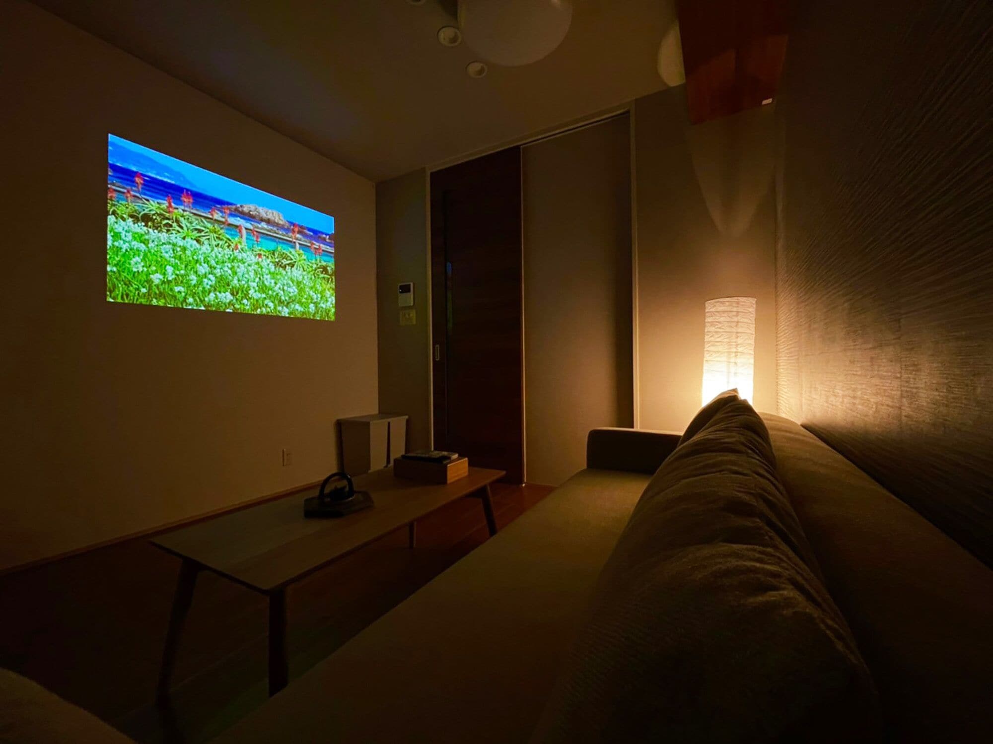 A dimly lit room with a large screen displaying a nature scene, a sofa, a small table, and a lamp.