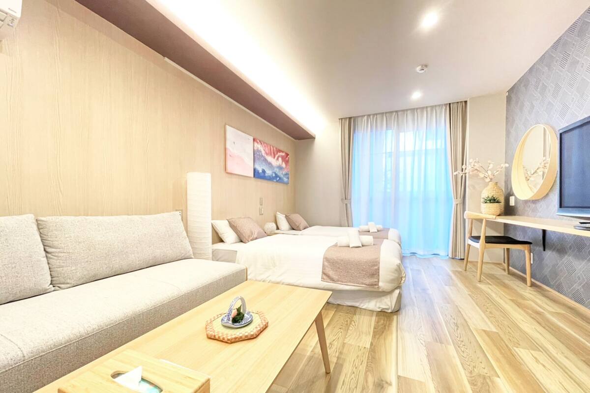 Una habitación de hotel moderna con sofá, dos camas, una mesita de madera, televisor y un amplio ventanal con cortinas.