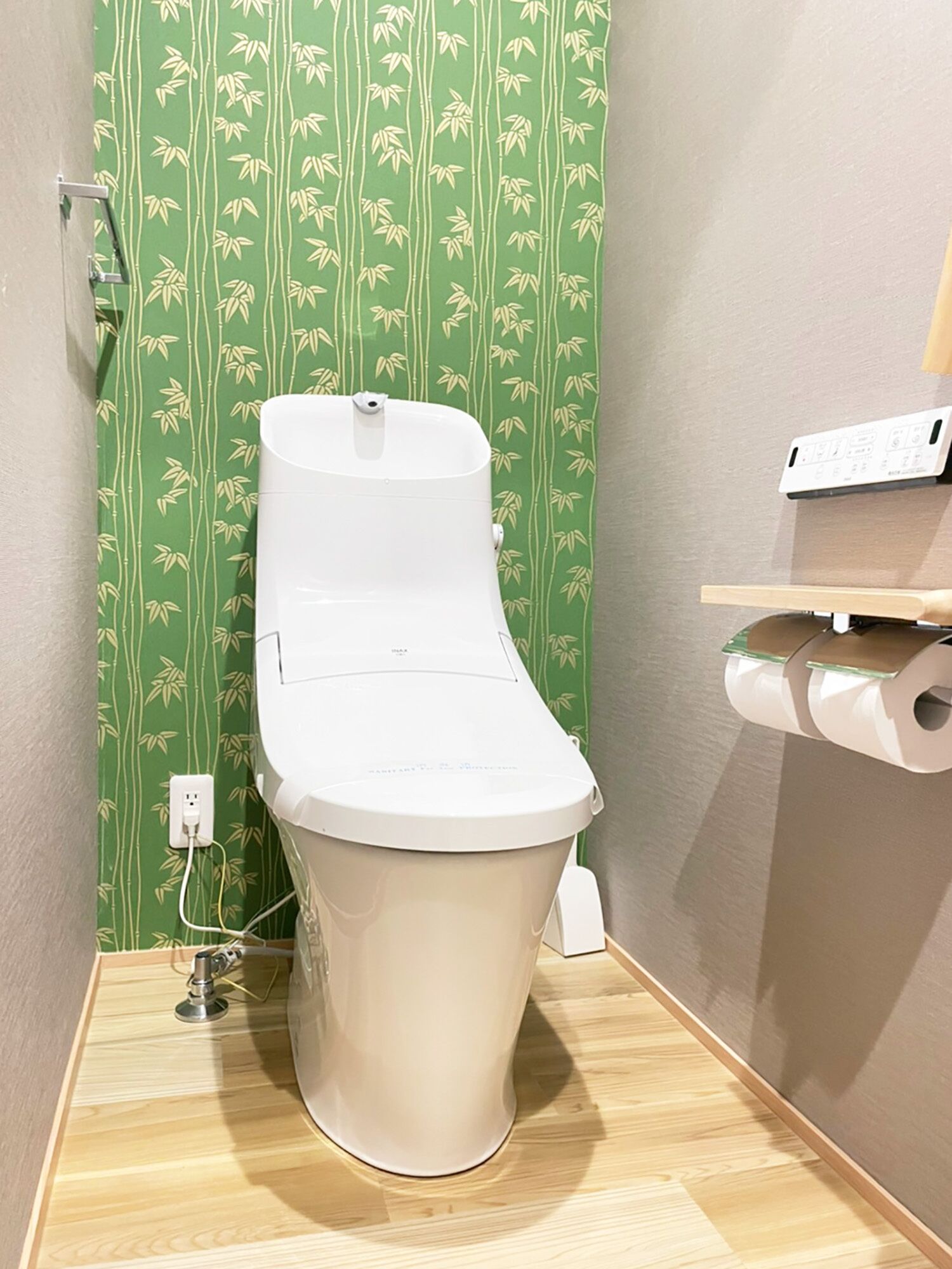 A toilet with a white seat and lid, a green patterned wall, a wooden floor, and a toilet paper holder on a shelf.
