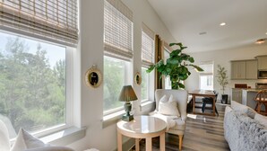 A spacious living area with a sofa, a round table, and a chair. There is a large window with blinds, a potted plant, and a view of trees outside.