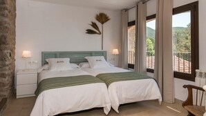 Ein Doppelbett mit weißer Bettwäsche und grünen Akzenten, eine Steinswand, ein Nachttisch mit Lampe und Blick ins Freie durch ein Fenster.