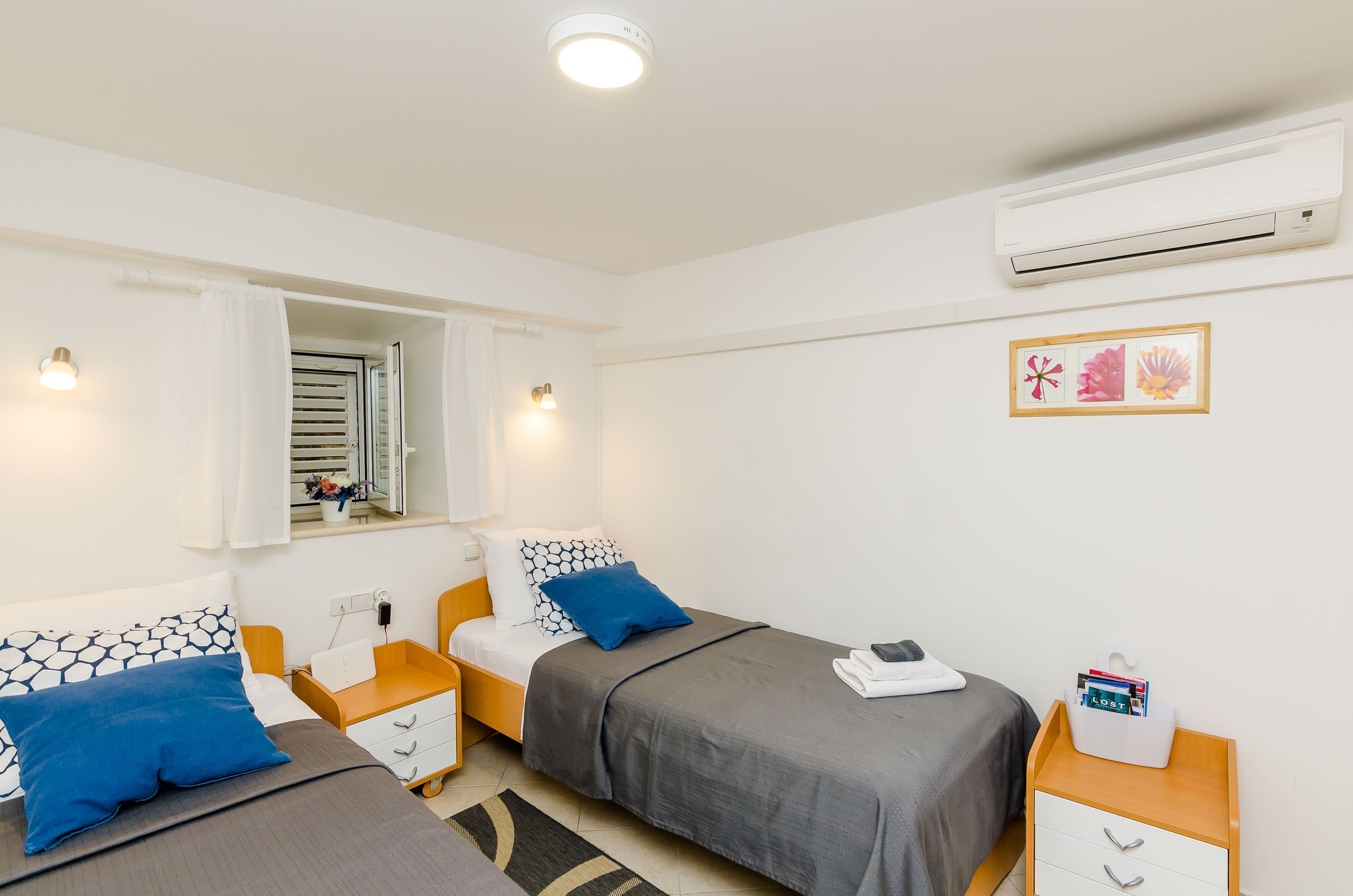 Ein Hotelzimmer mit zwei Betten, einem Fenster mit Vorhängen, einer Klimaanlage, einem Nachttisch mit Lampe und einem an der Wand hängenden Bild.