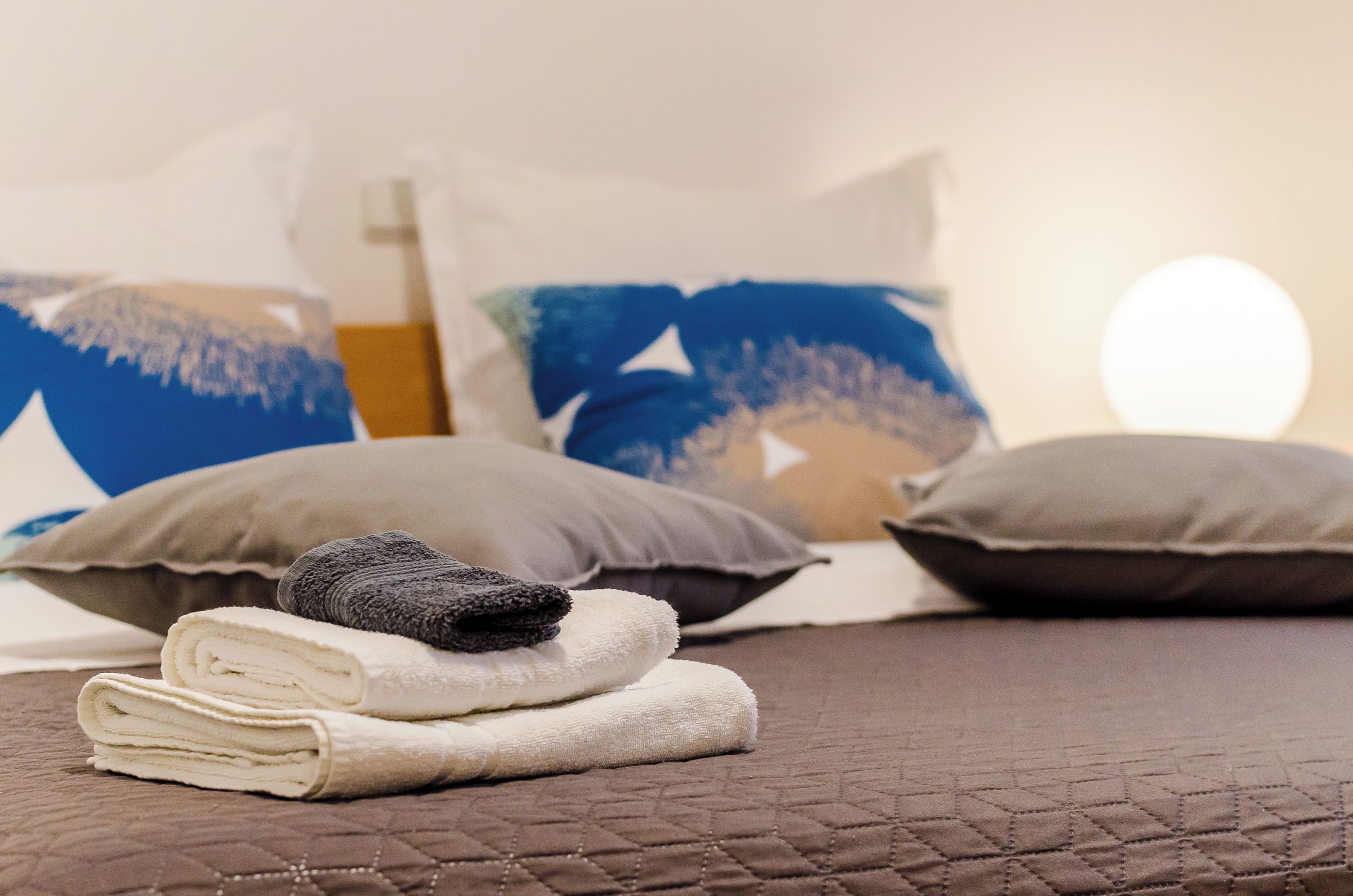 Una cama bien hecha con toallas enrolladas y almohadas, un edredón marrón acolchado, y una pared con una lámpara redonda.