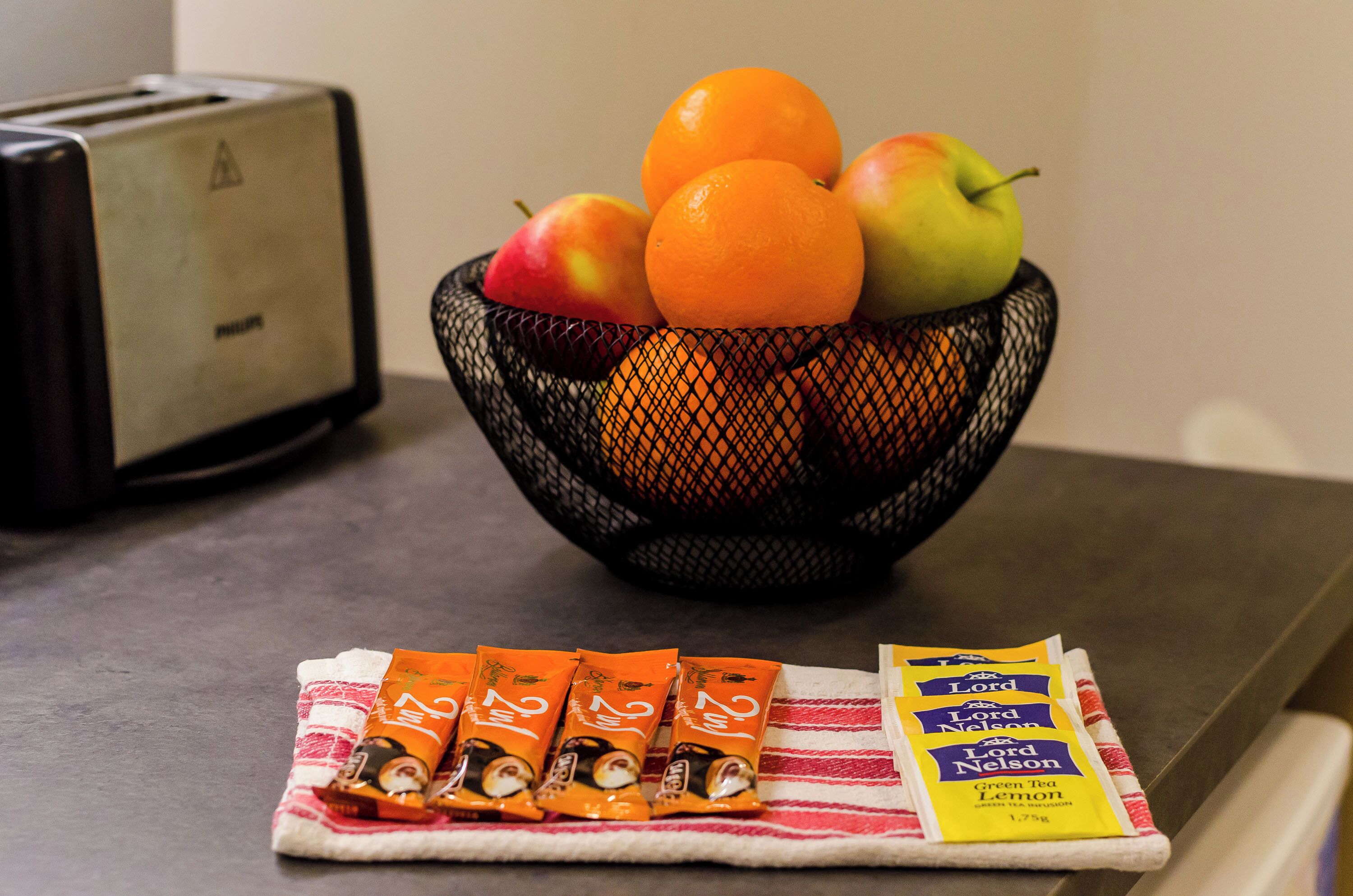 A fruit bowl with oranges and apples on a table, a toaster, and packets of tea on a cloth.