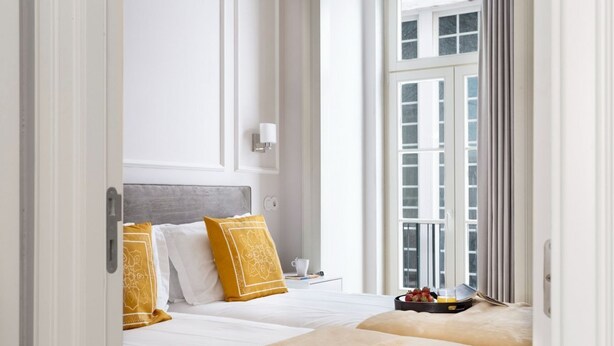 A hotel room with two beds, a bedside table with a cup and a tray of fruit, and a window with curtains.