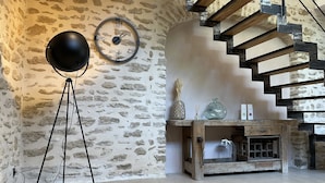 Habitació rústica amb paret de pedra, una làmpada de peu rodona, una taula consola de fusta amb objectes decoratius i unes escales de fusta.