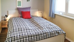 Siististi pedattu sänky, jossa on ruutukuviollinen päiväpeite, punainen tyyny ja lamppu yöpöydällä.