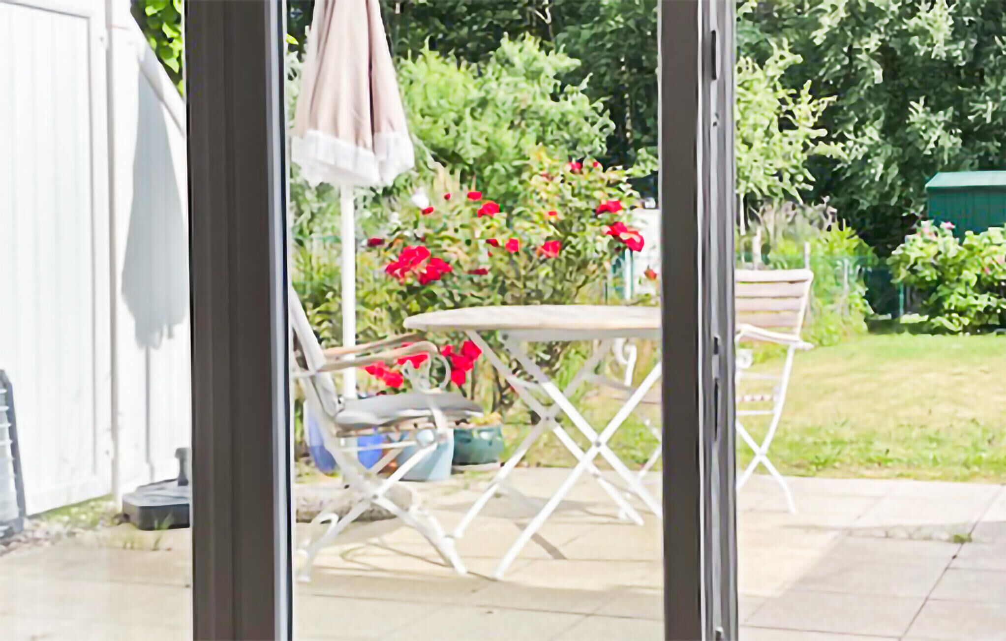 Une terrasse extérieure avec une table et des chaises, un parasol suspendu et un jardin agrémenté de fleurs rouges.