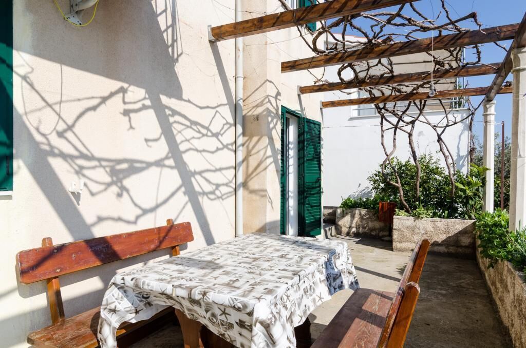 An outdoor patio with a table covered in a patterned tablecloth, a wooden bench, and a pergola with vines.