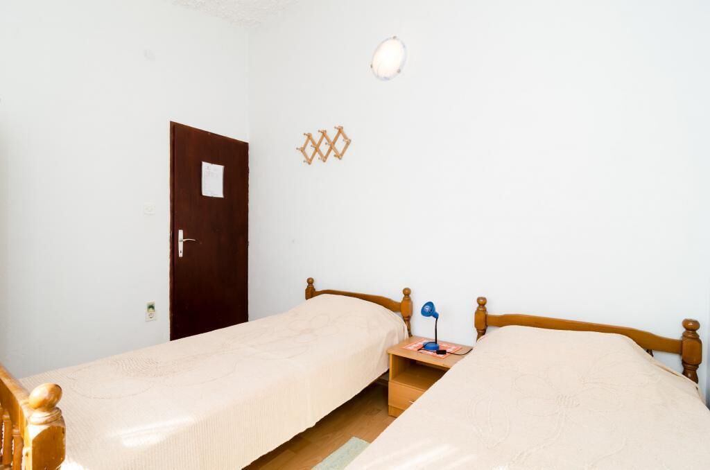 Una habitación pequeña y sencilla con dos camas individuales, una puerta de madera, una mesita con una lámpara y un aplique de pared.