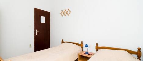 Una habitación pequeña y sencilla con dos camas individuales, una puerta de madera, una mesita con una lámpara y un aplique de pared.