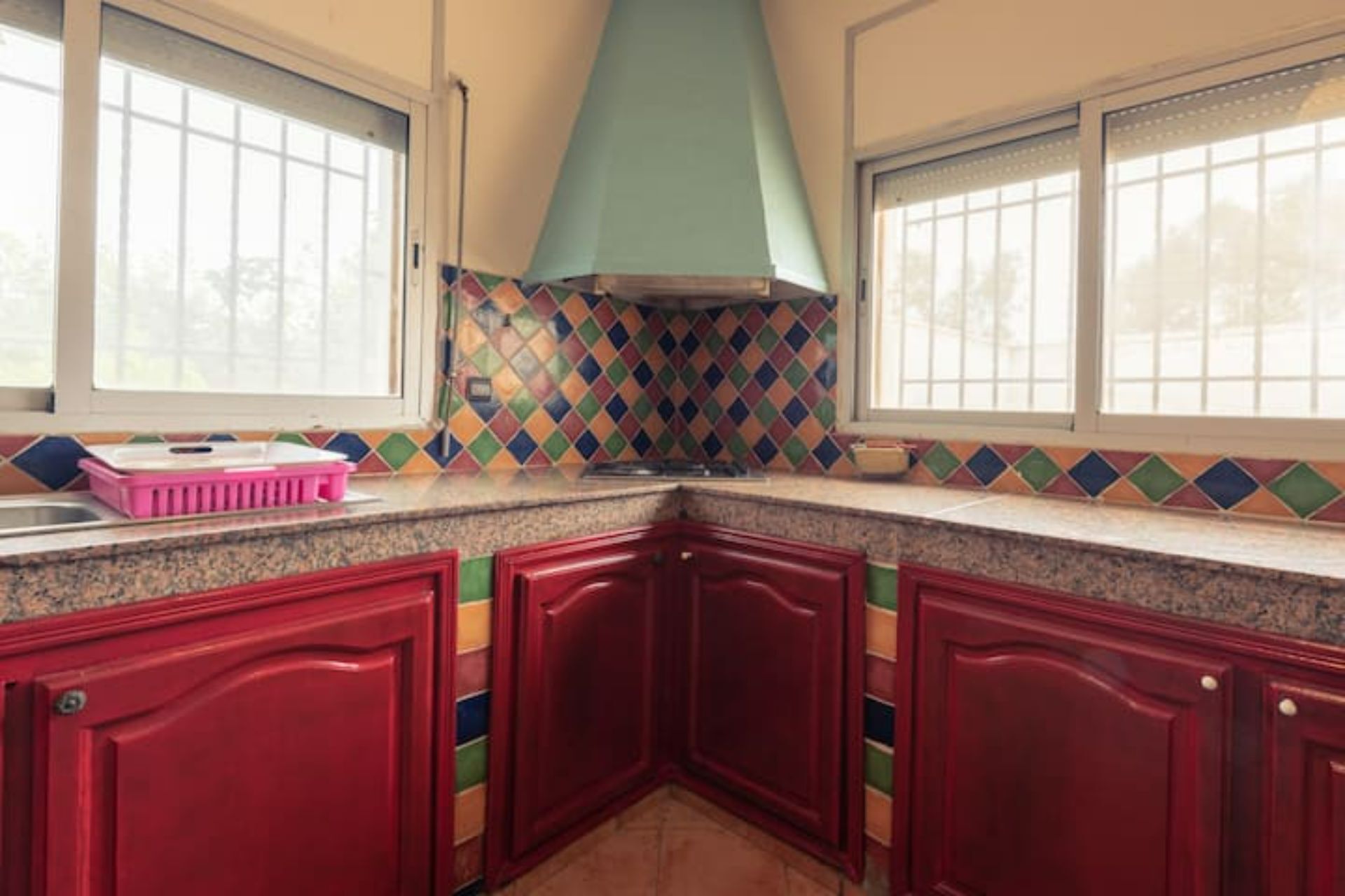 A kitchen with red cabinets, a colorful diamond-patterned backsplash, and a window with a white frame.