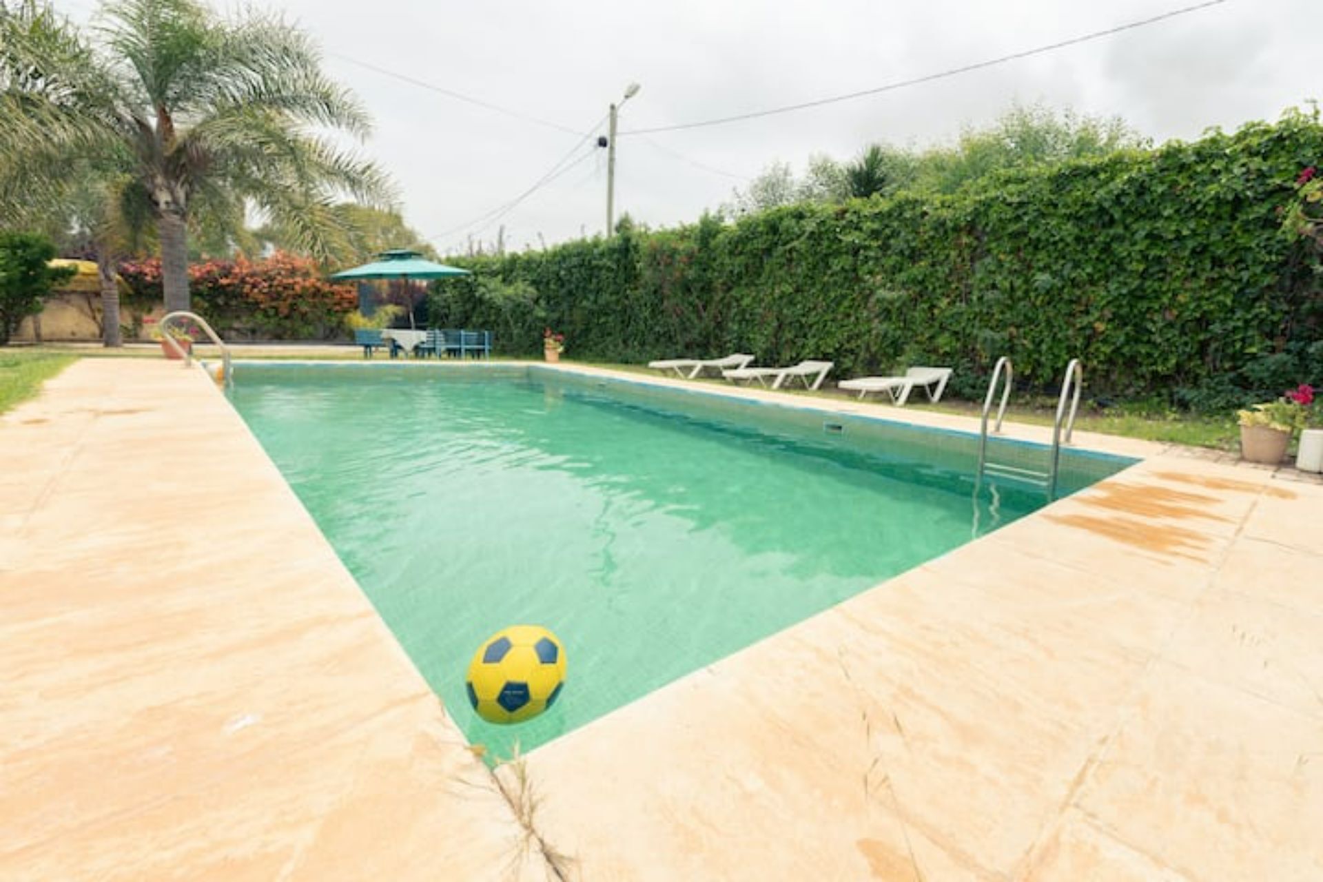 A rectangular swimming pool with a clear blue water surface, surrounded by a tiled deck. There are lounge chairs aligned along the pool's edge, and a single soccer ball is visible on the deck.