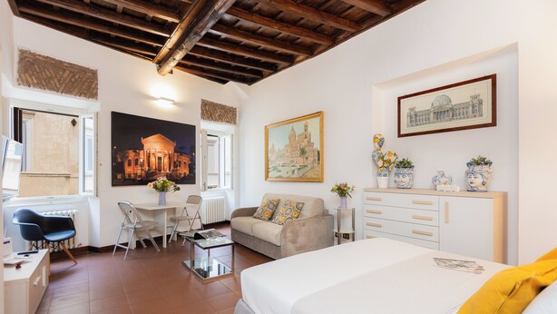 Une chambre d’hôtel moderne équipée d’un lit, d’un bureau, d’une chaise, d’un canapé et d’une table à manger. Les murs sont ornés de tableaux et il y a une fenêtre avec des stores.