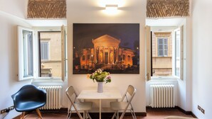 Une pièce comprenant une table à manger, deux chaises, un vase avec des fleurs et un grand tableau encadré représentant un bâtiment.