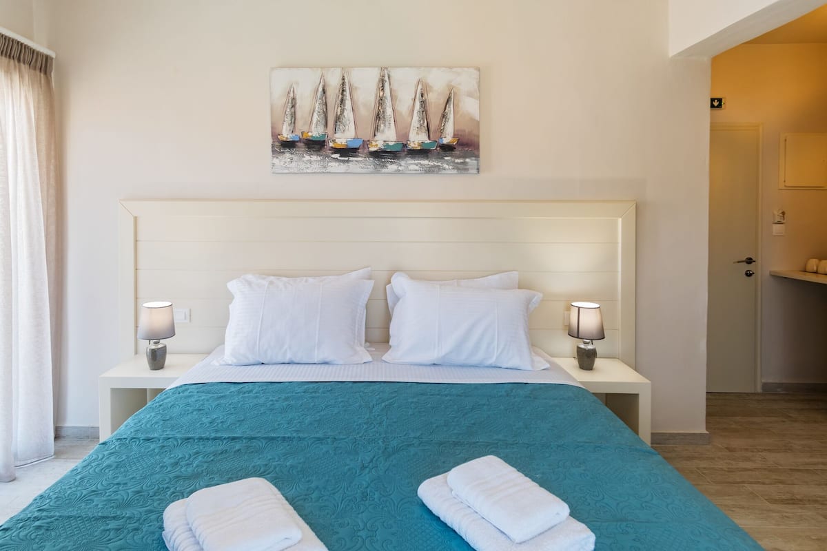 Una habitación de hotel con una cama grande, dos almohadas blancas, una colcha color turquesa, dos lámparas de noche y un cuadro de veleros en la pared.